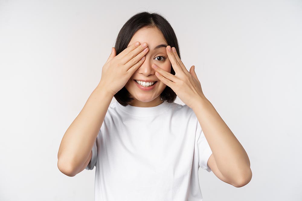 Portrait of asian woman covering eyes, waiting for surprise blindfolded, smiling and peeking at camera, standing over white background